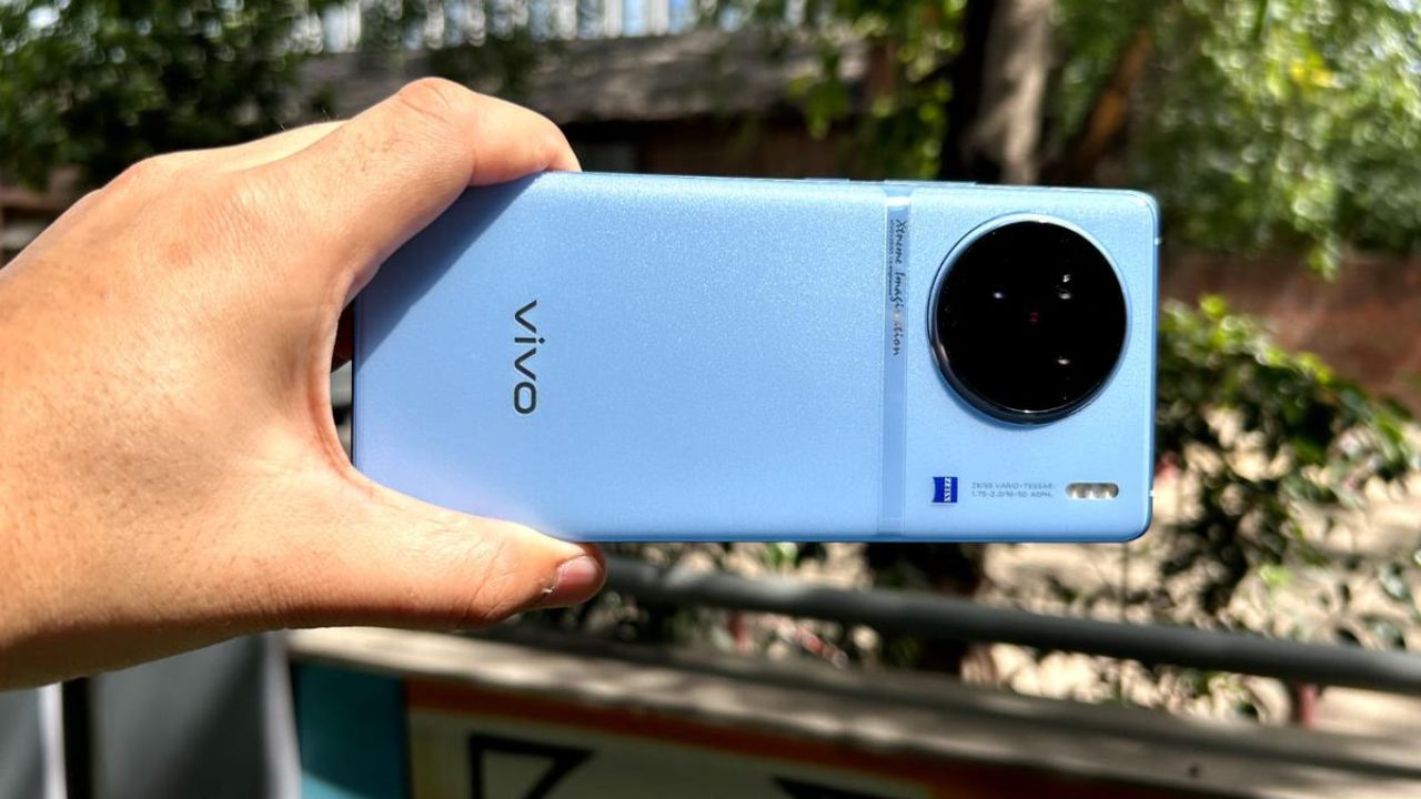 a man hold in in their hand vivo x90 in blue color in greenry background