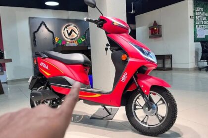 Here Is image of a man pointing a Elctric Scooter of Hero Electric Atria LX Which is in Red Colour and placed in showroom