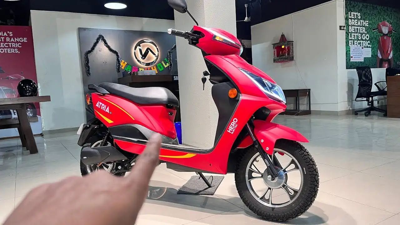 Here Is image of a man pointing a Elctric Scooter of Hero Electric Atria LX Which is in Red Colour and placed in showroom