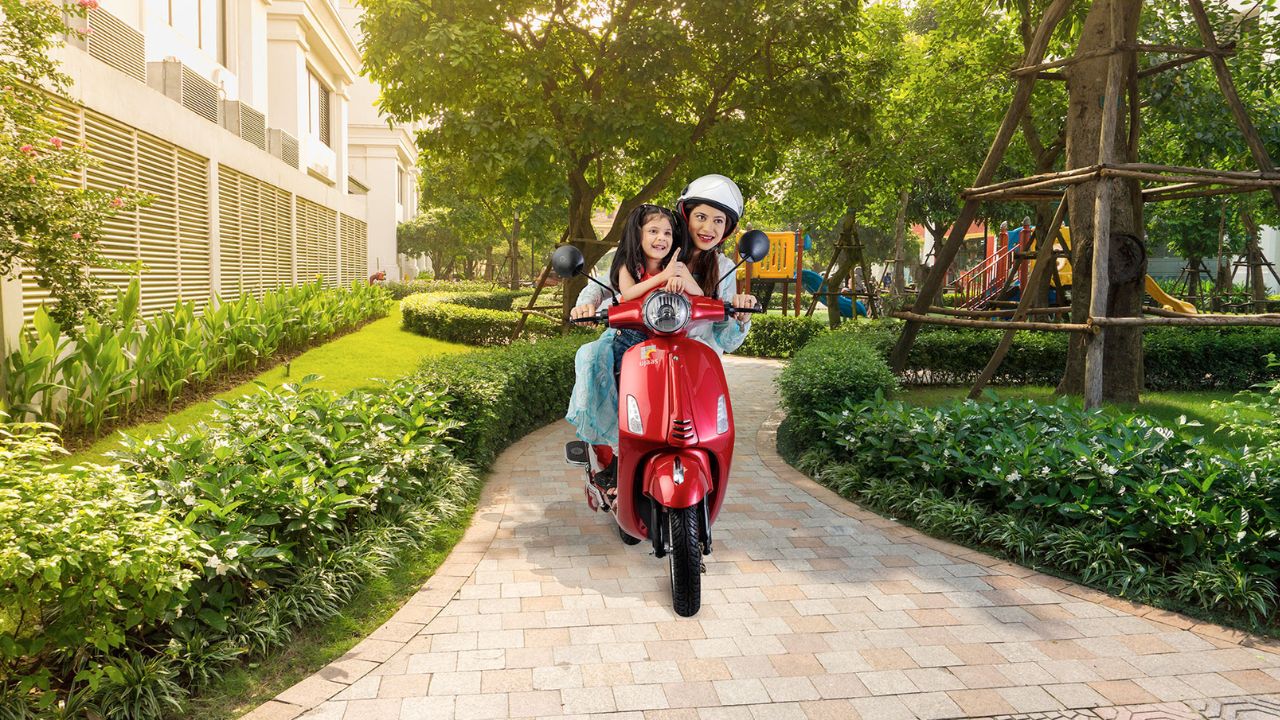 Here is image of A lady and a baby girl sitting on Ujaas Espa LA Electric Scooter which is placed in garden